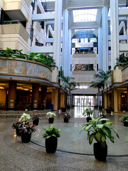 This image captures the interior of a modern building, possibly an office or corporate facility. The atrium is spacious and well-lit, featuring high ceilings with exposed beams and skylights. On the ground floor, you can see several potted plants arranged in patterns along the walkway. A few people are present in the space, going about their work or activities. There's a central courtyard with a glass ceiling where natural light filters through. The atrium is flanked by two levels of offices on either side, suggesting a multi-level office environment. Overall, the image conveys a sense of professionalism and contemporary design. DSC08454.JPG