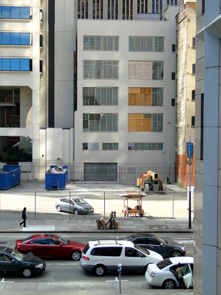 The image showcases a vibrant urban landscape. The scene is dominated by a large, industrial-style building with multiple windows and a prominent corner entrance. Adjacent to this building is a smaller structure, partially obscured from view. Both buildings are under construction or renovation, as indicated by the presence of scaffolding and protective barriers. In front of these buildings, there's a street that appears to be a one-way street, with cars parked along its sides. The surrounding area features typical city elements such as traffic lights and parking meters. This image captures a moment of everyday life in an American city. DSC08448.JPG