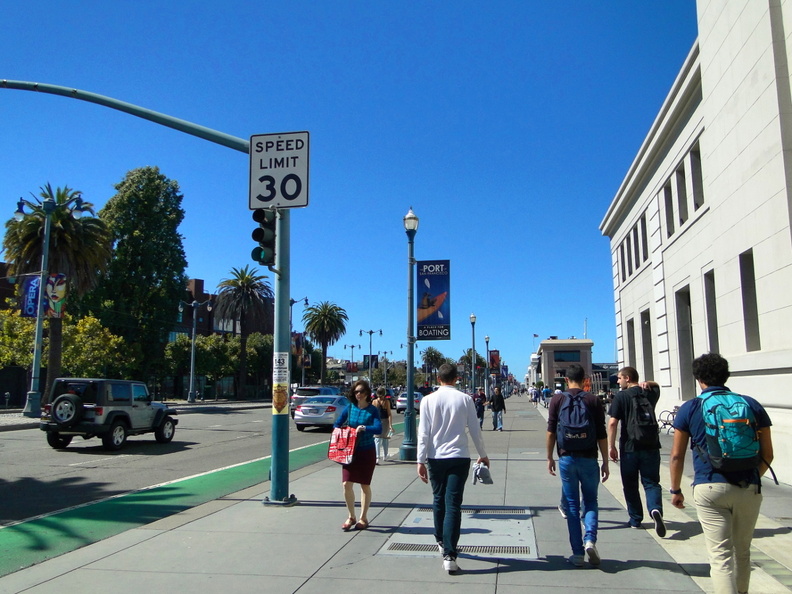 A sunny day in San Francisco, USA, showing a group of pedestrians walking down the sidewalk adjacent to a speed limit sign. The city scene includes various people, some carrying backpacks and handbags, walking along the street. Buildings can be seen in the background, suggesting an urban setting. DSC08568.JPG