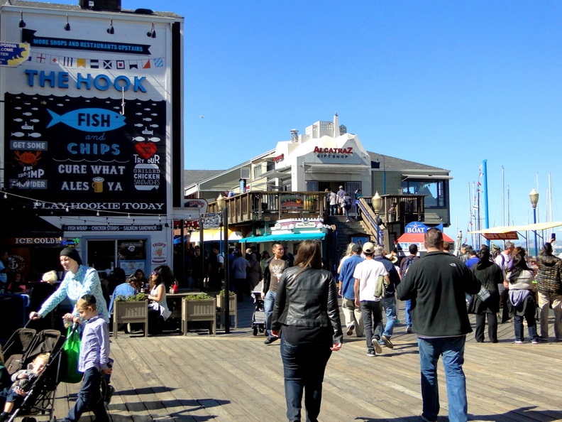 A bustling boardwalk with a lively atmosphere, featuring shops, restaurants, and people enjoying the outdoors. A sign for 'The Hook' is prominent, advertising fish and chips among other items. DSC08619.JPG