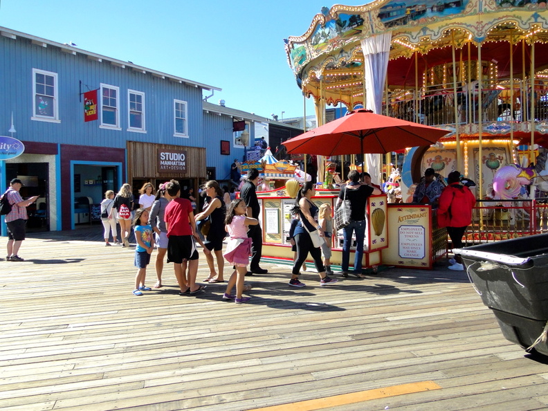 A bustling scene captures the joyful atmosphere of an amusement park. The focal point is a colorful carousel, complete with horses that are ready for riders. Surrounding the carousel is a lively crowd of people, some walking while others are engaged in various activities typical of such venues. A food stand, identifiable by its vibrant colors and umbrella, adds to the charm of the setting. The clear blue sky above suggests a pleasant day for outdoor fun. DSC08631.JPG