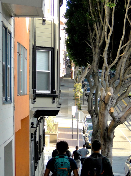 A photograph capturing a moment of two individuals walking down a tree-lined sidewalk in the city. They are casually dressed, one carrying a backpack, suggesting they might be tourists or commuters. The architecture and lush greenery suggest a well-maintained urban environment. DSC08909.JPG