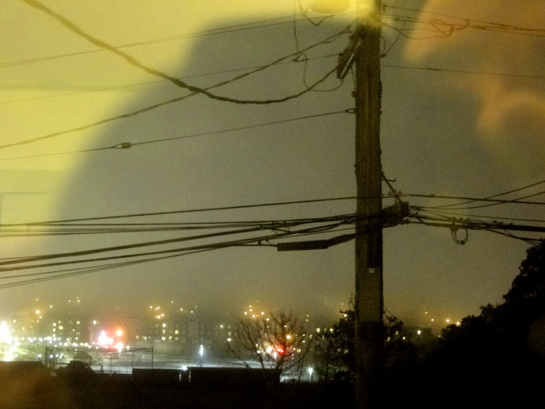 The image captures a city's nighttime ambiance during rain and fog. The scene is framed by power lines, with the glow of streetlights contrasting against the dark sky. A solitary figure can be seen in the window on the right side of the photo, adding a touch of human presence to the otherwise atmospheric urban landscape. DSC08986.JPG