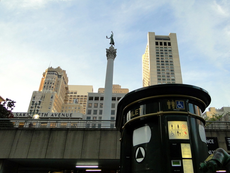 The image showcases a vibrant city scene, featuring a bustling street corner with a mix of historic and modern architecture. Dominating the view is a towering skyscraper, adorned with various signs and advertisements that hint at the urban character of San Francisco. The statue in the foreground adds an artistic touch to the cityscape, while the overcast sky casts a soft light over the scene. DSC08975.JPG