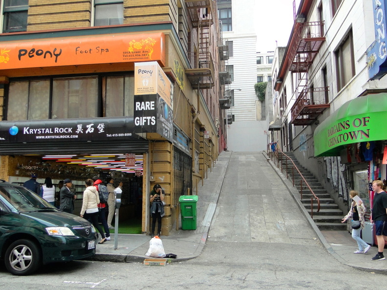 This image depicts a busy city street corner in San Francisco, showcasing the steep incline of the sidewalk and stairs. There are shops on both sides of the street, including one with a sign for 'Pony' and another called 'Rare Gifts'. The steep stairs are an iconic element in this urban environment. The area is bustling with pedestrians, who appear to be going about their day. A car can be seen parked along the sidewalk, and a truck is driving on the street. The buildings exhibit a classic San Francisco architectural style, with storefronts and signs that add character to the scene. DSC09066.JPG