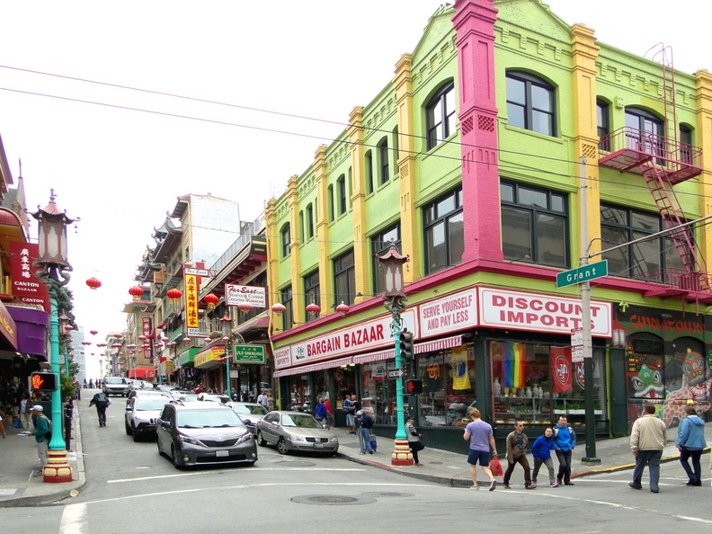 The image captures a lively scene from a bustling street in San Francisco's Chinatown. The street is flanked by colorful buildings, including a prominent pink and green building on the left side of the photo. Storefronts with signs and awnings line the sidewalk, inviting pedestrians to explore their offerings. People are seen walking along the sidewalk, adding life and energy to the scene. The street appears to be a popular spot for both locals and visitors, as indicated by the numerous cars parked or moving on the road. DSC09023.JPG