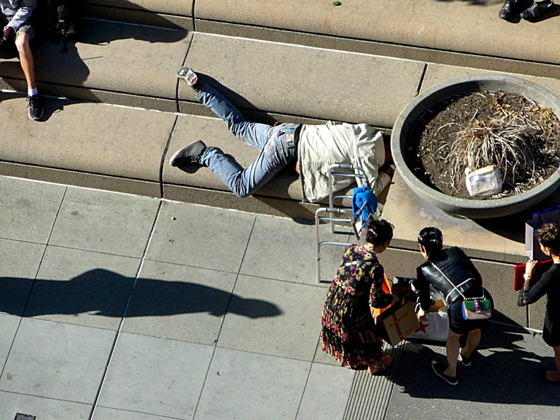 This image captures a poignant moment of a homeless man sleeping on a sidewalk in San Francisco. The scene is set in an urban environment, with pedestrians walking by and a person engaged in an act of kindness or concern by stopping to look at him. It's a stark reminder of the issue of homelessness in many cities. DSC09114.JPG