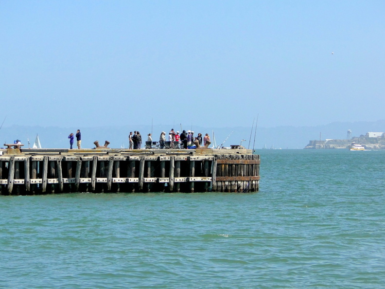 The image captures a serene moment at a fishing pier located near a body of water, possibly an ocean or a large bay. The pier extends into the distance, with a few people visible, likely engaging in recreational activities such as fishing or simply enjoying the view. In the background, there is a clear sky and what appears to be a bridge or structure on the far side of the water, which might suggest an urban setting in San Francisco. DSC09203.JPG