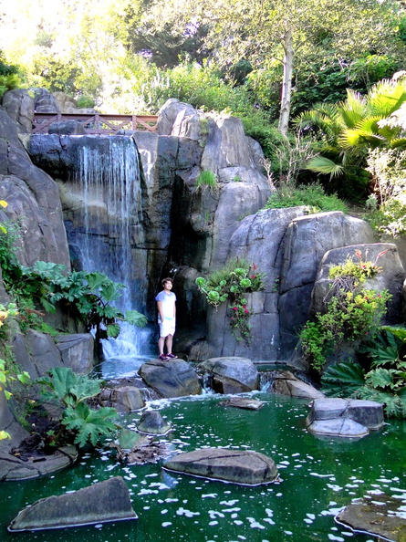 This image captures a tranquil scene in a park-like setting. A person is seen standing near the waterfall, perhaps enjoying the peaceful environment or taking a moment to appreciate nature. The area around the waterfall features lush greenery, providing a natural and serene atmosphere. The combination of stone elements, like the steps leading down to the waterfall, with the surrounding foliage adds a touch of rustic charm to this picturesque scene. It's a snapshot of a moment where one can escape from daily life and immerse themselves in the beauty of nature. DSC09372.JPG