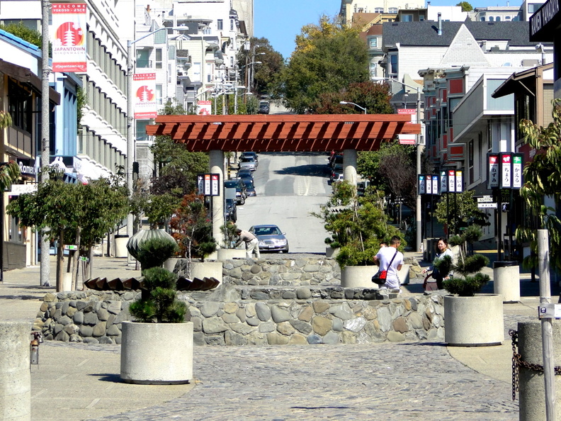 This image captures a typical day on a city street in San Francisco, USA. The scene includes a pedestrian walkway lined with various plants and trees, a brick pathway leading up to a small wooden canopy, and a parking lot adjacent to the street. There is a person seated on one of the benches under the canopy, enjoying the sunny day. In the distance, buildings and hills can be seen, indicating that this area is part of the city's neighborhood. The sky is clear, suggesting pleasant weather. DSC09441.JPG