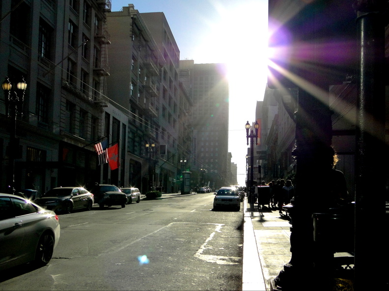 This image captures a serene moment in the heart of San Francisco, California. The sun shines brightly, casting long shadows and illuminating the scene with warm, natural light. A one-way street stretches out into the distance, lined by buildings and dotted with parked cars. In the foreground, a lone pedestrian adds a touch of life to the urban landscape. Above, an American flag flutters gently on a nearby building, adding a patriotic touch to this snapshot of city life. DSC09512.JPG