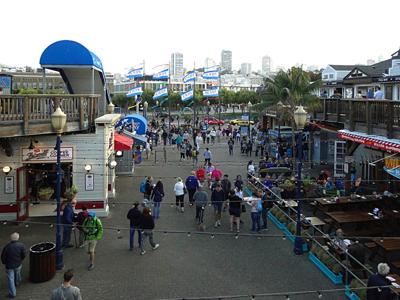 The image captures a lively city festival, bustling with people walking around the venue. The open area is filled with attendees and various event tents, indicating a vibrant atmosphere. In the background, you can see a street lined with shops and restaurants, typical of a downtown area in a large city. The sky appears to be overcast, suggesting the possibility of rain during the event. Overall, this scene conveys a sense of community and celebration. DSC09517.JPG