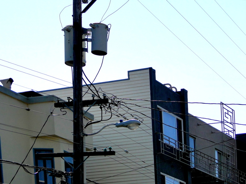 The image showcases a typical residential street in San Francisco, characterized by its dense infrastructure. Dominating the scene is a utility pole and various power lines crisscrossing above, indicative of a city where technology plays a vital role in daily life. DSC09477.JPG