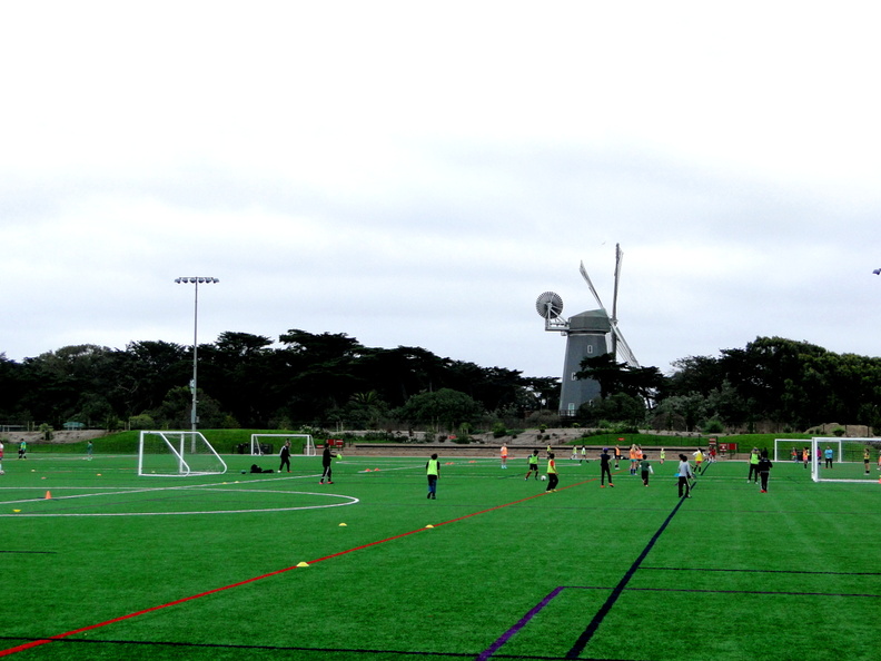 A lively soccer match is taking place on a sunny day, with players actively engaged in the game. The goalkeeper, dressed in yellow, stands alert behind the goal, ready to intercept any incoming shots. The field is surrounded by green grass and a solitary windmill can be seen in the distance, adding to the picturesque setting. DSC09942.JPG