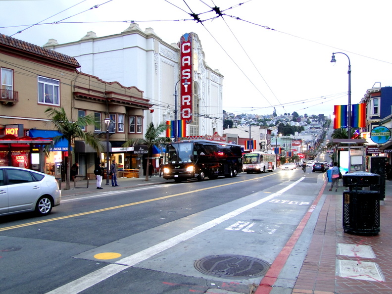The image captures a vibrant city scene in San Francisco, California during what appears to be rainfall. The street is lined with colorful buildings and businesses, including the Castro Theatre. Vehicles are navigating the wet roadway under overcast skies. A few pedestrians can be seen as well, going about their day despite the weather. The presence of traffic lights and a city bus suggest an active public transportation system. DSC09959.JPG