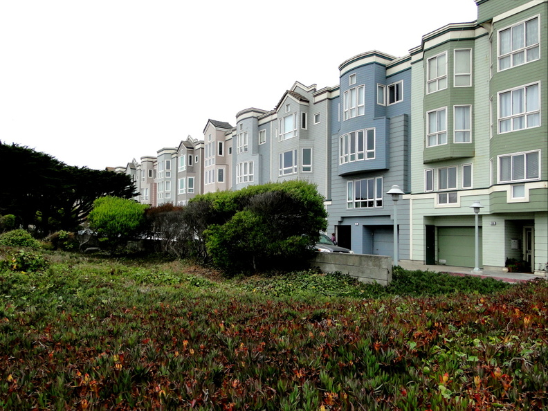 The image depicts a residential area in San Francisco, with a row of multi-story apartment buildings. The facades are consistent, suggesting it might be a gated community or an organized neighborhood. There's a lush green lawn separating the buildings from what appears to be a park or public space. The architecture is characteristic of American cities, with clean lines and modern design elements. DSC09898.JPG