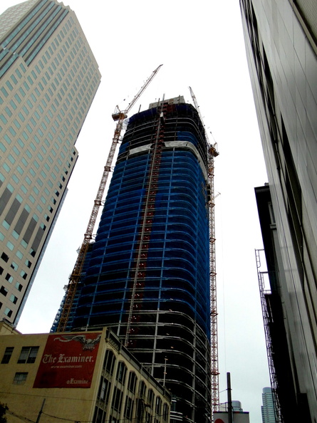 This image captures a vibrant city scene, featuring an under-construction high-rise building dominating the downtown skyline. The construction crane stands tall above the partially completed structure, signifying ongoing development in the urban landscape. In the foreground, a glimpse of daily life can be seen with a pedestrian and a store front, while the background provides a panoramic view of other city buildings and structures. DSC09864.JPG