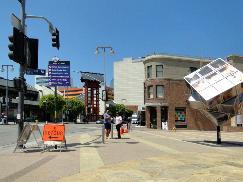 A vibrant cityscape with a striking piece of public art located at an intersection. The art appears to be a large, reflective sculpture shaped like a letter 'M', which adds a unique aesthetic and landmark to the street corner. Surrounding the sculpture is a sidewalk bustling with pedestrians, traffic lights regulating the flow of vehicles, and street signs indicating directions or local businesses. DSC07966.JPG