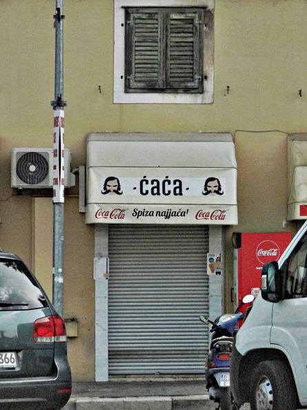 The image features a local cafe named 'Cafe' with the signage displaying both 'Caca' and 'Spana'. The shopfront is situated on the ground floor of a building. In front of the store, there is a closed metal shutter. A car is parked next to the cafe. The style of the photograph suggests it may have been taken for commercial or real estate purposes. DSC02308.JPG