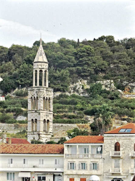 The image depicts a stone church with a tall bell tower located in the heart of an old town. The architecture suggests it might be a historical Roman Catholic church. The setting includes buildings typical of a coastal Mediterranean town, and the surrounding landscape indicates that this town is situated near the sea. DSC02331.JPG