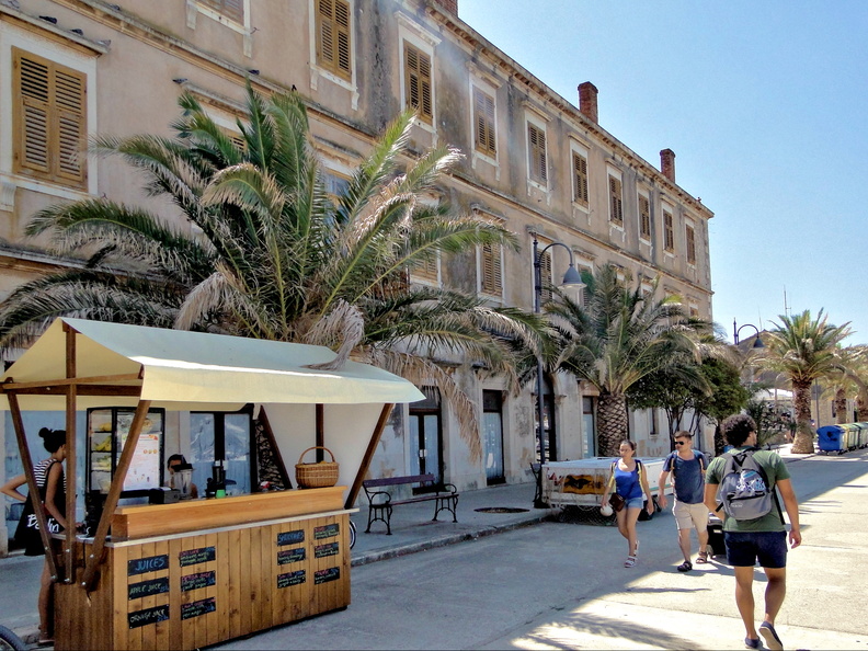 The image captures a vibrant street market in Stari Grad, Croatia. A small, thatched-roof stall is set up on the sidewalk, offering shade and inviting passersby with its inviting display of goods. The market is nestled within an old town, characterized by historical buildings and palm trees lining the streets, adding a touch of Mediterranean charm to the scene. DSC02857.JPG