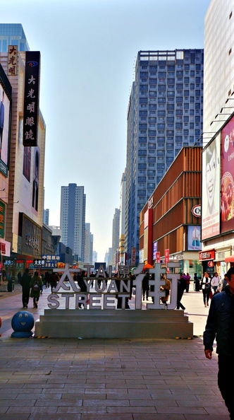 This image captures the bustling atmosphere of Shenyang, China. The street is alive with activity; numerous pedestrians are going about their day on the sidewalks and the overhead signage suggests a variety of shops and businesses catering to the city's residents and visitors. In the foreground, there is a large banner advertisement for a movie, adding a touch of color and excitement to the urban landscape. The sky is clear with no visible clouds, indicating good weather conditions at the time of capture. This scene is typical of many Chinese cities where street-side vendors, signage in both Chinese and English, and pedestrian traffic are common features. DSC07231.JPG