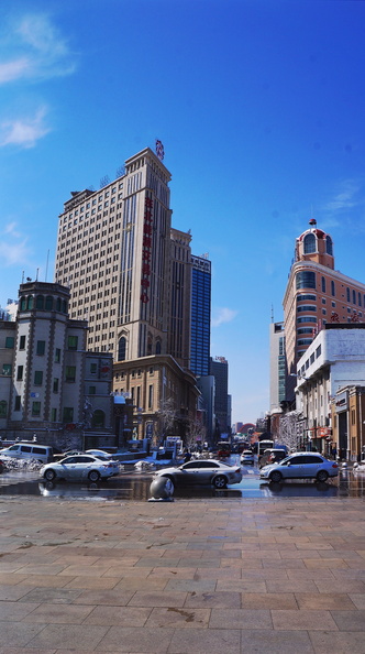 The image depicts a bustling city street corner in the heart of Shenyang, China. The perspective is from street level looking up at the impressive skyline, where modern skyscrapers tower over historical buildings, creating a stark contrast between old and new architectural styles. The sunny day illuminates the cityscape with a clear blue sky, which adds vibrancy to the urban environment. DSC07334.JPG