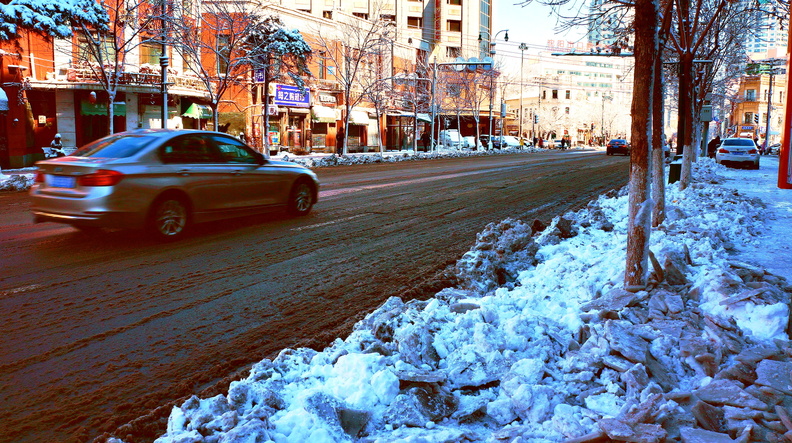 The image captures a winter day in a city. A car is seen driving down the snow-covered street, which appears to be a main road given its length and lack of obstacles. The street is lined with trees that have been stripped of their leaves by the cold weather, adding to the seasonal ambiance. On the right side of the image, a sidewalk is visible with a small amount of snow piled up on the left side. Buildings are in the background, suggesting this scene is located within an urban environment. DSC07343.JPG
