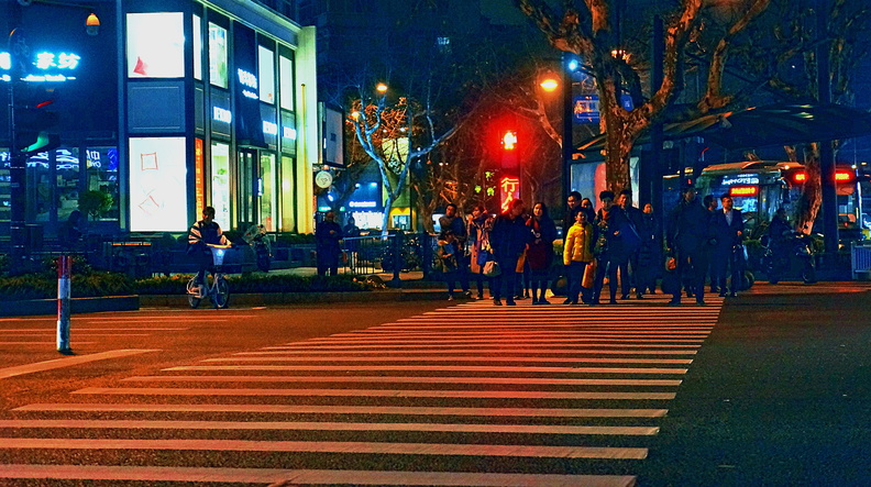 This image captures the lively atmosphere of a nighttime scene in Hangzhou, China. The intersection is bathed in streetlights and vehicle headlights, creating a dynamic contrast between light and shadow. People can be seen crossing at the crosswalk, while vehicles are parked along the street. A red traffic light stands out against the city lights, indicating that this moment was captured during the evening rush hour. DSC07436.JPG