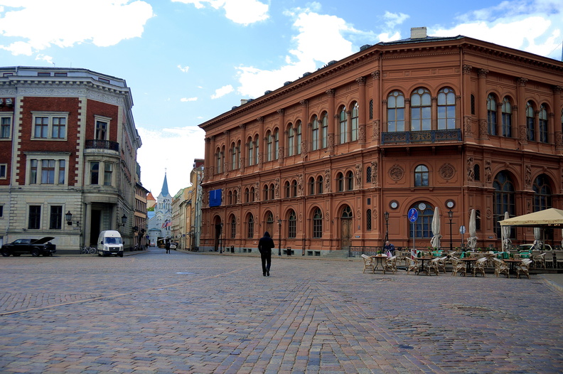 A picturesque scene from the heart of Riga, Latvia. This image captures the beauty and charm of the city's historic district, with its well-preserved buildings made of brick and stone, and a cobblestone street that adds a touch of old-world elegance. The clear day allows for an unobstructed view of the surroundings, showcasing the blend of traditional and modern elements. Visible on the street are tables and chairs, perhaps belonging to outdoor cafes or restaurants, inviting passersby to sit and enjoy the atmosphere. Pedestrians can be seen walking along the paved courtyard, going about their day in this bustling part of town. DSC09847.JPG