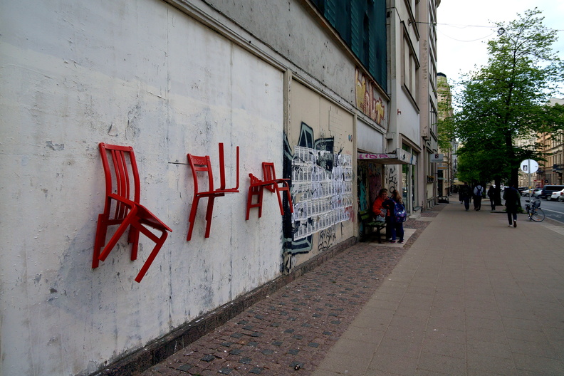 This image captures a vibrant scene on the streets of Riga, Latvia. The street corner is adorned with a colorful mural and features four red chairs mounted on a wall. The chairs, painted in a striking shade of red, are arranged casually as if inviting passersby to sit and enjoy the local art. In the background, we see a typical European cityscape with buildings that add depth to the image. The overall atmosphere is one of creativity and community engagement. DSC09976.JPG