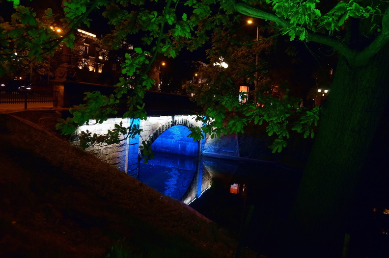 This image captures the tranquil beauty of an urban area during the evening hours. A stone bridge with a low arch spans over a calm river, illuminated by soft streetlights that cast a gentle glow on the water surface below. The bridge's reflection shimmers on the water, adding to the peaceful ambiance. The surrounding area is lush with trees and foliage, creating a natural contrast against the man-made structures of the cityscape. The overall scene conveys a sense of solitude and quietness, as if time has momentarily paused in this picturesque locale. DSC00424.JPG