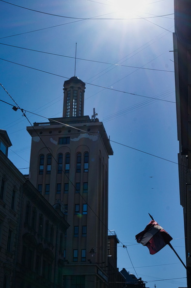 The image depicts a sunny day in Riga, Latvia, showcasing the Art Nouveau Central Market building along Riga's famous Kalnu Street. The focal point is a tall clock tower, visible above other buildings. The clear blue sky provides ample sunlight that illuminates the architectural details of the tower and the surrounding area. Power lines crisscross above the street, indicating an urban setting. A flag can be seen on the right side of the image, possibly on a flagpole within the frame. The overall scene conveys a sense of calm and everyday city life. DSC00009.jpg