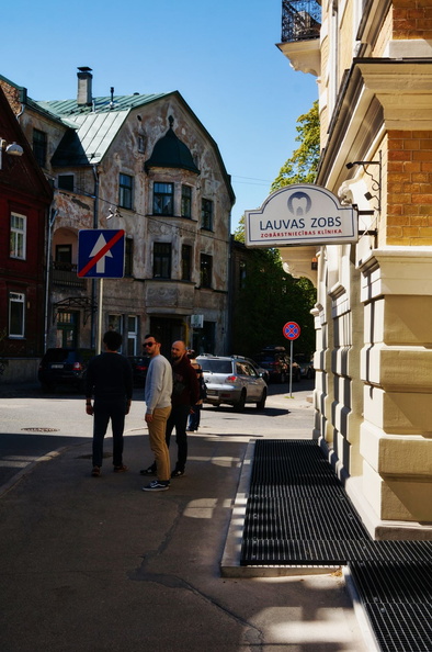 This image captures a quiet moment on a residential street in Riga. A blue sky forms a backdrop to the white building, which is adorned with a sign. The street itself is lined with buildings and trees, adding a touch of greenery to the urban landscape. People can be seen leisurely strolling along the sidewalk, enjoying the pleasant weather. The overall scene is representative of everyday life in a European city. DSC00189.jpg