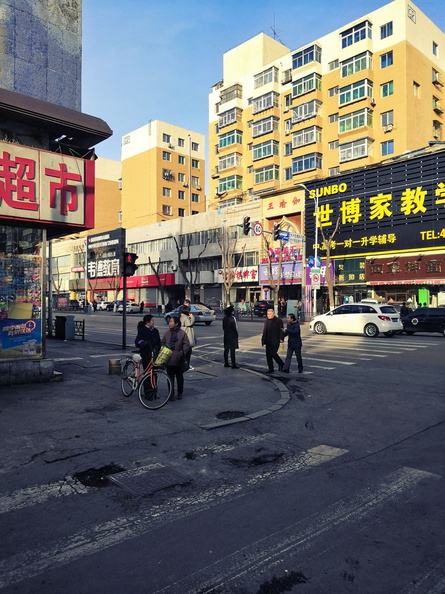 This image captures a bustling street in Shenyang, China. The scene is filled with people going about their day, walking along the sidewalks and crossing the streets. The presence of colorful signs, typical of Chinese urban landscapes, adds to the lively atmosphere. The buildings, likely shops or small businesses, line the street, and the weather appears pleasant, inviting locals and tourists alike to explore this vibrant city. IMG_20180321_161455.jpg