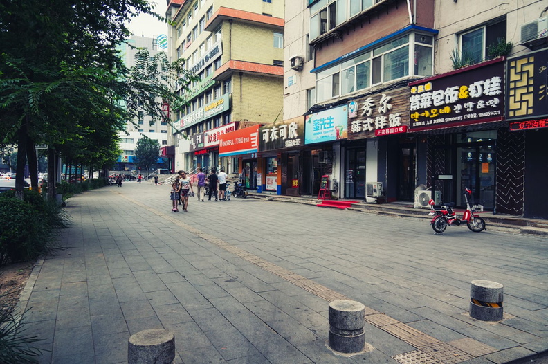 This image captures a bustling street scene in Shenyang, China. The walkway is lined with various shops and restaurants, as indicated by the signage. There are several people enjoying the outdoors on this sunny day. Trees provide a touch of greenery amidst the urban landscape. A bicycle parked along the path adds to the everyday life charm of this cityscape. DSC00949.jpg