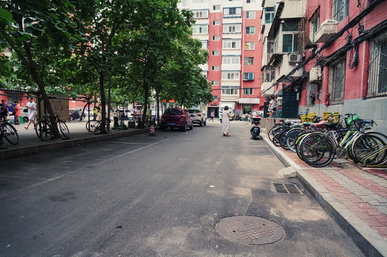 This image captures a typical day in an urban residential area. The street is lined with trees and buildings, showing signs of daily life with bicycles parked along the side. There's a car parked on the left side of the road, and a few people can be seen going about their day. DSC00953.jpg