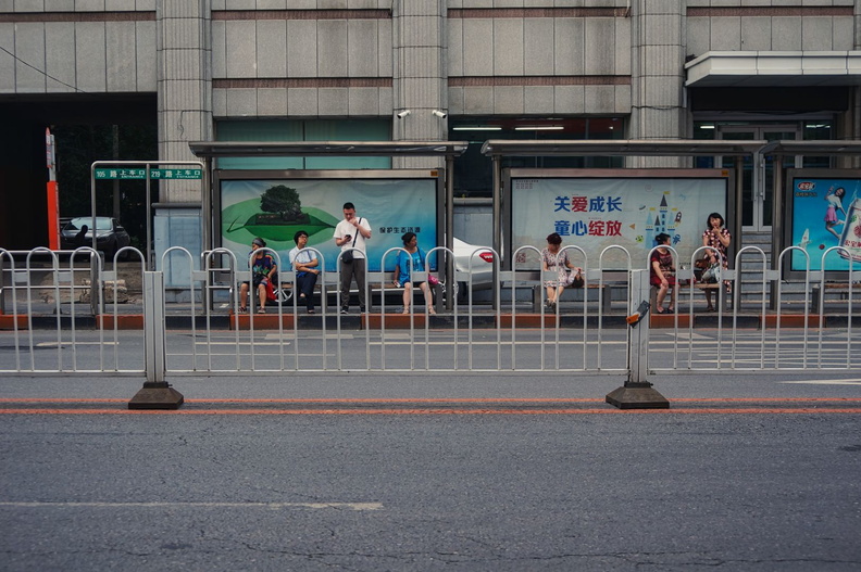 This image captures a moment in urban China, featuring a bustling bus stop with people waiting to board the bus. The bus stop is located on the street corner, where you can see pedestrians, public transportation, and the vibrant city life. The architecture of the buildings in the background suggests a modern city setting. In this scene, there are several elements that make it representative of everyday life in China. DSC00973.jpg