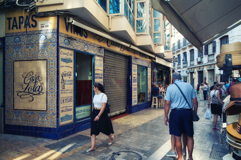 This image captures a lively street scene in the heart of Malaga, Spain. The vibrant atmosphere is palpable with locals and tourists alike strolling along the sidewalks. A prominent feature of the street is a charming blue-and-white tiled facade with Spanish writing, which adds to the European charm. Shops and cafeterias line the streets, inviting passersby to explore their offerings. The clear sky above suggests a pleasant day for visitors. DSC01105.jpg
