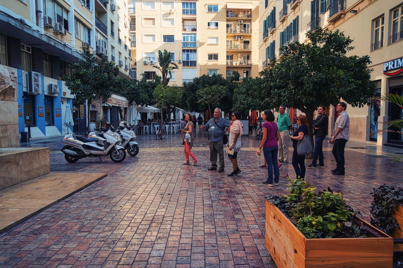 This image captures an outdoor gathering space on the rooftop of a residential building in Malaga, Spain. The area is paved with large stone blocks and features a variety of seating options, including benches and chairs. There are trees providing shade and adding to the inviting ambiance. The scene takes place during dusk, as indicated by the soft, warm lighting that enhances the textures and colors in the space. DSC01145.jpg