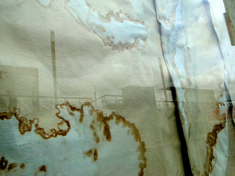 A French window with a rusted curtain overlooking the sea and industrial structures in the background.